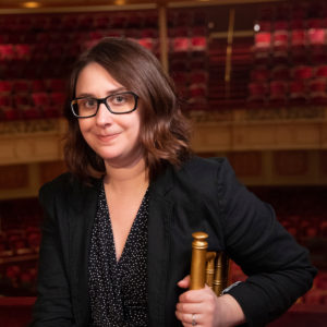 Shanna is a white woman with medium length brown hair and large black eyeglasses. She sits in the opera box at the Carnegie Music Hall positioned on the 2nd floor above the stage, overlooking the audience.