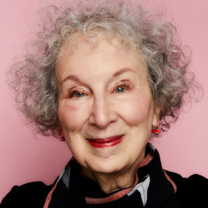 Headshot of Margaret Atwood with a pink background. Atwood is a senior white woman wearing a beautiful scarf and red lipstick, with short curly hair. She smiles into the camera.